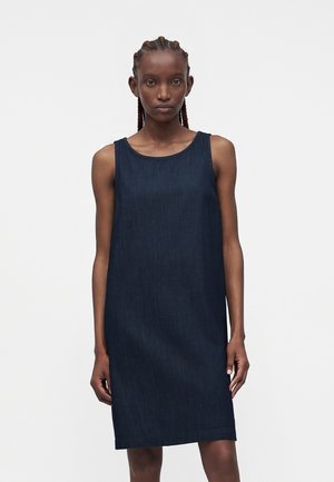 Γυναίκα με πλεξούδες, ντυμένη με μαν sleeveless μίντι, σκούρο μπλε φόρεμα από τζιν, μπροστά σε απλό λευκό φόντο.