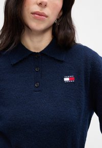 Námornícky modrá pletená polo košeľa s rebrovaným golierom, troma čiernymi gombíkmi a štvorcovou nášivkou s nápisom „Tommy“ v bielej a červenej farbe.