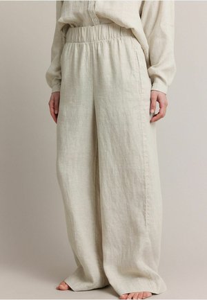 Luchtige beige linnen broek met elastische taille, gedragen door een persoon die blootsvoets staat, handen rustend langs de zij, met een bijpassend beige overhemd aan.