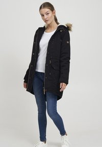 Black parka jacket with fur-lined hood, pockets, and zip closure. Worn over a white shirt and blue jeans, paired with white sneakers.