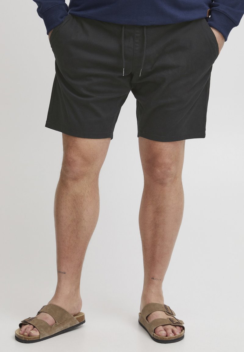 Pantalones cortos negros con cintura elástica y cordón, que cuentan con bolsillos laterales; combinados con sandalias marrones que presentan un detalle de hebilla.