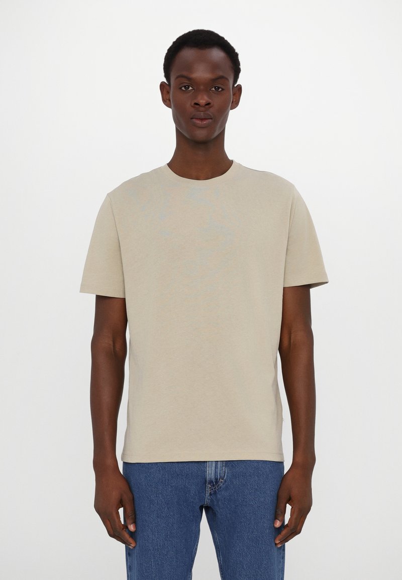 Camiseta de algodón beige de mangas cortas y cuello redondo, combinada con jeans de mezclilla azules. Diseño simple, textura ligera, sin patrones visibles.