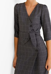 Robe à carreaux gris avec un design portefeuille, des manches trois-quarts, une ceinture à nouer et deux boutons sur le devant. Tissu texturé avec un motif subtil.