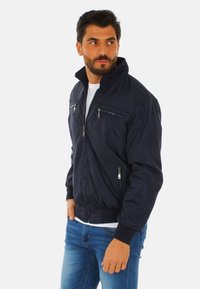 Giacca bomber blu navy con chiusura a zip, due tasche sul petto e polsini a costine. Tessuto leggero con una texture liscia e design classico.