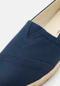 Scarpa slip-on in tela blu navy con tessuto texture e dettagli in cucitura; suola in corda di juta con forma arrotondata e caratteristiche di design minimal.