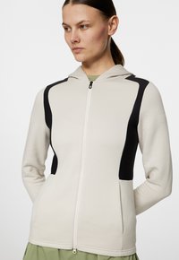 Ljust grå zip-up hoodie med svarta sidopaneler, texturerad tyg och huva. Har en framdragkedja och fickor. Slät yta.