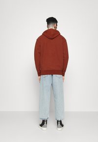 Sudadera con capucha de color óxido y corte relajado, combinada con jeans de pierna ancha en azul claro y zapatillas negras de caña alta.