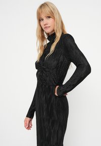 Fekete, hosszú ujjú ruha álló gallérral, függőleges redőkkel és csavart részlettel a deréknál. Testhez simuló fazon.