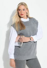 Suéter gris de punto sin mangas sobre una camisa blanca de botones, combinado con leggings grises. Ella sostiene un bolso metálico y lleva pendientes de aro.