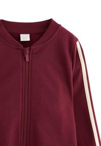Bordeaux-farvet zip-up jakke med ribbet halsudskæring, der har et cremegult lodret strib langs ærmet. Fremstillet af blødt stof med en glat tekstur.