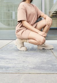 Brąsowa, oversize sukienka T-shirt z miękkiego materiału, zestawiona z jasnobeżowymi, masywnymi platformami i wzorzystymi skarpetkami do kostek, umieszczona na kamiennej podłodze.