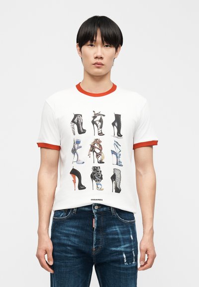 Νέος άνδρας που φοράει λευκό T-shirt με κόκκινο περιθώριο, στο οποίο απεικονίζονται εννέα διαφορετικά σχέδια ψηλοτάκουνα παπούτσια, και μπλε τζιν, σε απλό φόντο.