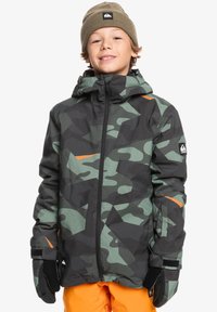 Vattentät jacka i kamouflagemönster med svart och grönt och orange detaljer. Har huva och textilstruktur. Handskar och orange byxor synliga.