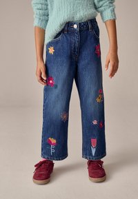 Jeans de mezclilla de pierna ancha, con bordados florales coloridos en varias formas. Acompañados de zapatos de ante rojos. Suéter azul claro.