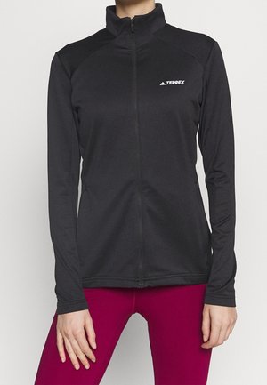 Nainen pukeutuneena mustaan täysvetoketjulliseen Terrex-takkiin, jossa on pitkät hihat, ja magenta-värisiin leggingseihin, seisomassa yksivärisen taustan edessä.