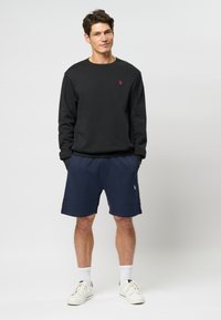 Fekete kapucnis pulóver kis piros logóval, navy színű rövidnadrággal párosítva, lazán illeszkedő fazonban, elasztikus derékrésszel, fehér sportcipővel viselve.