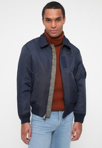 Veste bombardier bleu marine avec une doublure matelassée grise, des poignets côtelés et une fermeture à zip. Elle est dotée d'une poche sur la poitrine et de poches latérales, portée par-dessus un col roulé rouille.