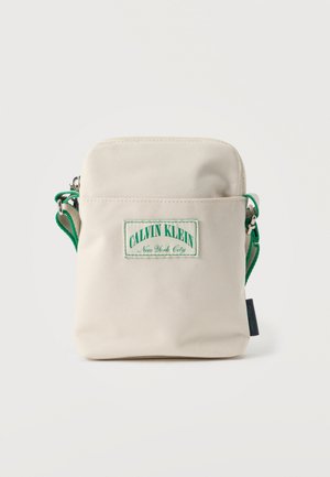 Calvin Klein beige fabric crossbody bag with green strap and embossed logo patch. Compact rectangular shape with zip closure and side label.