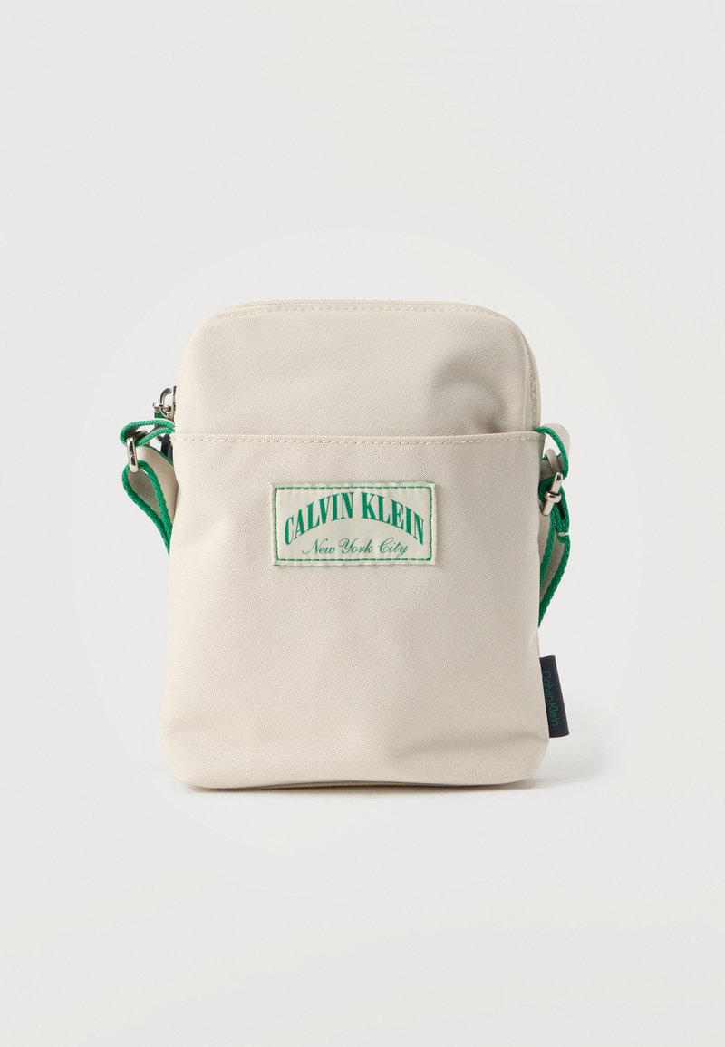 Μπεζ υφασμάτινη τσάντα Calvin Klein χιαστί με πράσινο λουράκι και ανάγλυφη ετικέτα με λογότυπο. Συμπαγής ορθογώνιο σχήμα με φερμουάρ στο πάνω μέρος και πλαϊνή ετικέτα.