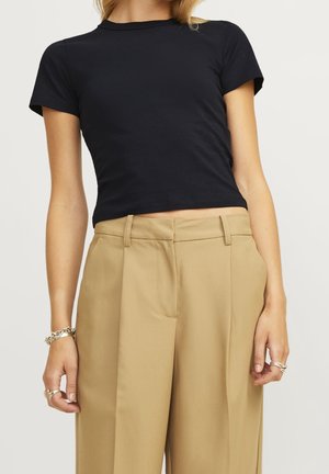 Femme portant un crop top noir ajusté à manches courtes et un pantalon beige à taille haute et jambes larges, accessoirisée avec des bagues et un bracelet.