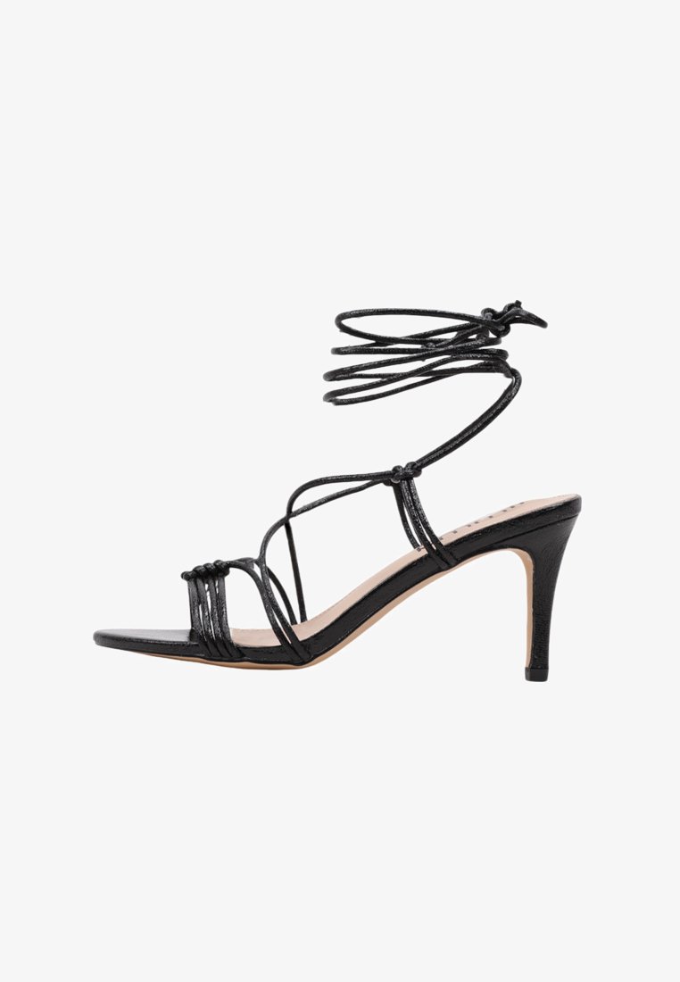 Sandalia de tacón alto negra con tiras finas, diseño entrecruzado y lazo en el tobillo. La suela es lisa y el tacón es afilado.