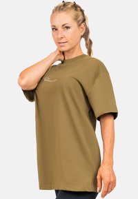 T-shirt oversize verde oliva, maniche corte, collo rotondo, con un logo ricamato discreto sul davanti. Tessuto liscio, vestibilità casual.