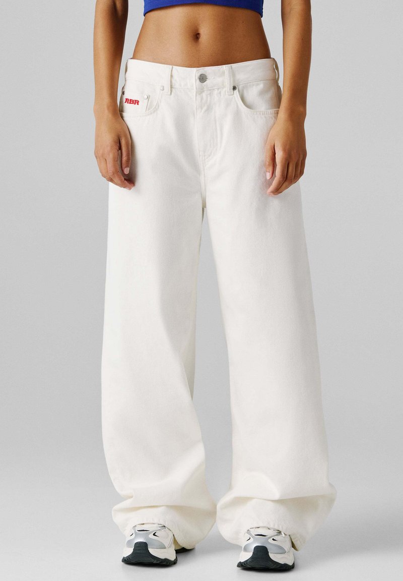 Henkilö pukeutunut ylisuuruihin valkoisiin farkkuihin, joissa punainen logo taskussa, siniseen crop-topiin ja valkoisiin lenkkareihin yksinkertaisen harmaan taustan edessä.