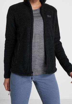 Person wearing a black zip-up fleece jacket over a gray shirt and blue pants, standing with arms relaxed by their sides.