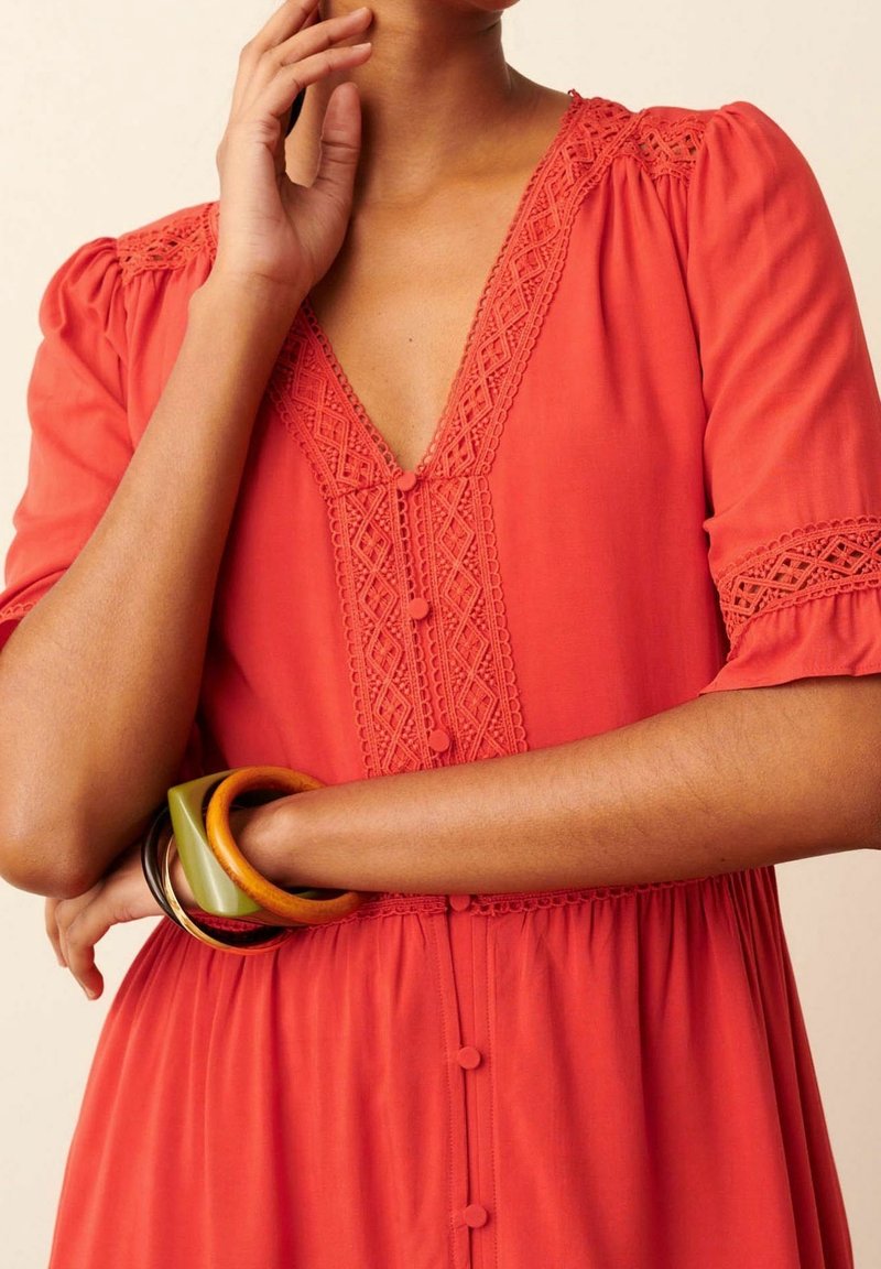 Femme portant une robe rouge avec des détails en dentelle et des boutons recouverts de tissu, accessoirisée avec des bracelets verts, marron et en bois sur le bras.