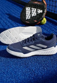Спортен маратонка Adidas в тъмносин и бял цвят на син корт за тенис с падъл ракета и тенис топка на заден план.