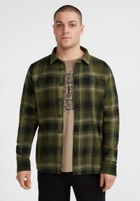 Groen en zwart geruit flanellen overhemd met een button-down kraag, met twee borstzakken, gedragen over een tan kleurig t-shirt met een graphic.