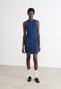 Denim brezrokavnik, prilagojena silhueta, temno modra barva, sprednji sredinski šiv, dolžina do kolen, v kombinaciji z belimi nogavicami in črnimi čevlji.