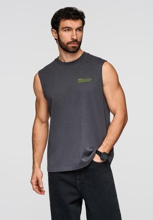 Mężczyzna z brodą ubrany w bezrękawnik w ciemnoszarym kolorze z małym żółtym napisem, czarne dżinsy i czarny zegarek, patrzący na bok z ręką w kieszeni.