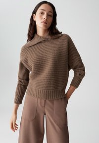 Bruine geribbelde gebreide trui met een opgerolde kraag en lange mouwen, gecombineerd met beige wijde broekspijpen. Zachte textuur, losse pasvorm.