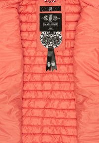 Rosa quiltad jacka med en inredning som har horisontella stygn, märkesetiketter och ett dekorativt svart band med detaljer. Mjuk, glansig textur.