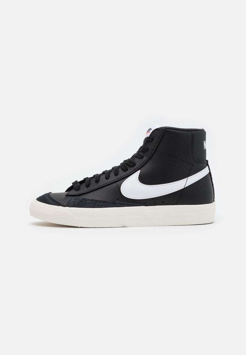 Korkeavartiset tennarit, jotka on valmistettu mustasta nahasta, valkoisella Nike-logolla, teksturoiduilla mustilla yksityiskohdilla ja vaaleankeltaisella kumipohjalla. Varustettu mustilla kengännauhoilla.