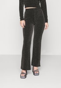 Svarta glittrande byxor med en utsvängd design, hög midja och slät yta, ihop med klumpiga blockklack-sandaler.