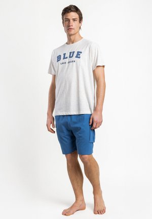 Joven de pie descalzo con una camiseta blanca con la inscripción "BLUE LOIS JEANS" y pantalones cortos cargo azules sobre un fondo blanco.