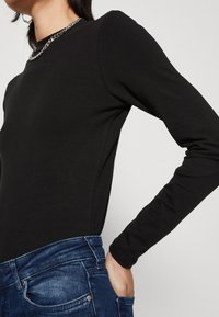 Schwarzes Langarmoberteil aus glattem Stoff, mit Rundhalsausschnitt und silberner Kettenakzent, kombiniert mit dunklen Jeans.