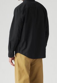 Schwarzes langärmeliges Hemd aus glattem Stoff mit geknöpftem Bündchen. Das Hemd hat einen abgerundeten Saum und wird mit khakifarbenen Hosen kombiniert.