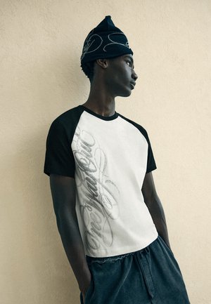 Jonge man draagt een zwart-wit t-shirt met geschreven tekst, donkere broek en een zwarte gebreide muts, staand tegen een beige gestructureerde muur.