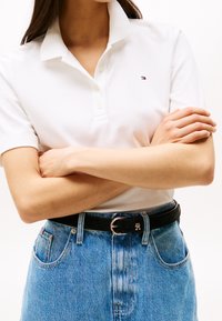 Personne portant un polo blanc avec un petit logo et un jean bleu, les bras croisés, et une ceinture noire avec une boucle argentée.