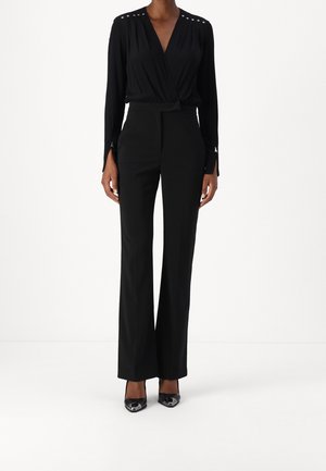 Kobieta ubrana w czarną, zapinaną na zakładkę bluzkę z długim rękawem ozdobioną srebrnymi ćwiekami oraz obcisłe czarne spodnie, do tego czarne, szpiczaste szpilki.