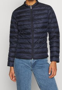 Person wearing a navy blue quilted zip-up jacket paired with light blue denim jeans, standing against a plain gray background.