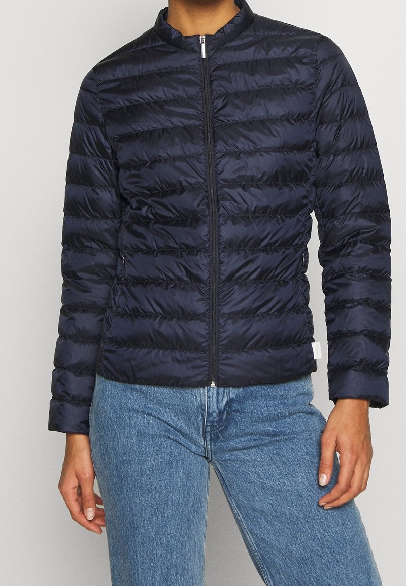 Person wearing a navy blue quilted zip-up jacket paired with light blue denim jeans, standing against a plain gray background.