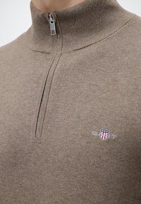 Pull en maille beige avec une courte fermeture éclair et un petit logo brodé représentant des étoiles et des rayures sur la poitrine.