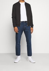 Černá bomber bunda, bílý tričko s kulatým výstřihem, modré rovné džíny, bílé tenisky, nošené s bílými ponožkami. Jednoduchý, neformální outfit.