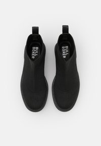 Botines negros de tobillo hechos de un tejido elástico con una textura suave. Cuentan con tiradores y una suela de goma. Logo visible en la plantilla.