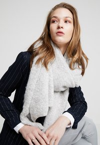 Foulard doux et léger de couleur gris clair, avec une finition texturée, drapé lâchement autour du cou, complétant un blazer rayé bleu et une chemise blanche.