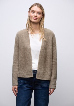 Młoda kobieta o blond włosach nosi beżowy, puchaty kardigan, biały top i ciemnoniebieskie dżinsy, stoi na neutralnym, jasnym tle.